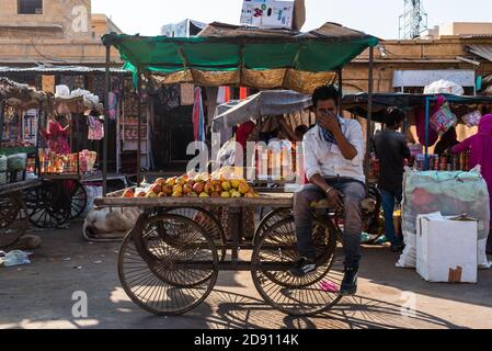 Jaisalmer, Rajasthan / Indien - november 02 2020 : Junge Erwachsene Mann fixiert seine Gesichtsmaske sitzt auf seinem eigenen Obst-Handwagen Verkauf von Früchten Stockfoto