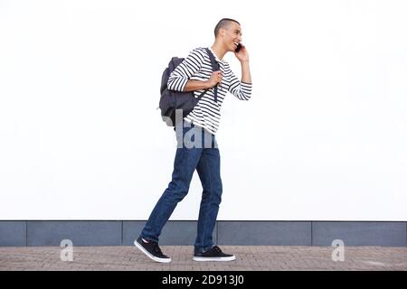 In voller Länge Seite Porträt von glücklich jungen Mann zu Fuß mit Tasche und Gespräch auf Handy gegen weiße Wand Stockfoto