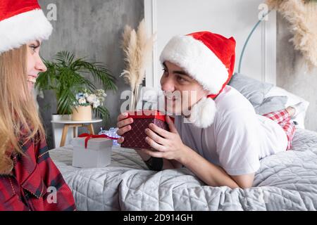 Menschen, Geschenke und Urlaub Konzept. Kaukasisches Paar zu Hause. Junger lächelnder Mann, der auf dem Bett liegt und Geschenkbox hält und auf glückliche Frau schaut, die auf Th sitzt Stockfoto