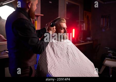 Selbstbewusster kaukasischer Barbier Mster schneidet Haare und Bart von Männern im Friseurladen, professioneller Friseur macht Frisur für einen jungen Mann, er arbeitet mit Haarschneidemaschine Stockfoto