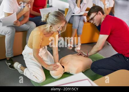 Junge kaukasische weibliche Ausbilderin zeigt, wie man Mann während des erste-Hilfe-Trainings in Innenräumen legt, isoliert im Zimmer mit einer Gruppe von Menschen Stockfoto