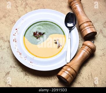 Traditionelles Küchenkonzept. Spinat- und Kürbiscremesuppe auf grauem Texturhintergrund. Japanische Suppe im Restaurant. Püree in grüner und gelber Farbe in Yin Yang Zeichen durch Pfefferbox, Salzkeller und Löffel Stockfoto