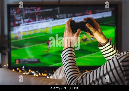 Nahaufnahme männliche Hände spielen Videospiel Fußball auf Konsole. Gamer Mann mit Gamepad Controller Halten Wireless Joystick sitzt auf Pouf in der Nähe Stockfoto