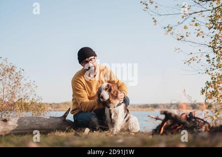 Der Mann umarmt seinen Hund, der am See sitzt. Aktive Erholung mit Haustieren, auf Abenteuer gehen, Gesellschaft: spaniel Hund und sein Besitzer sitzen im Freien Stockfoto