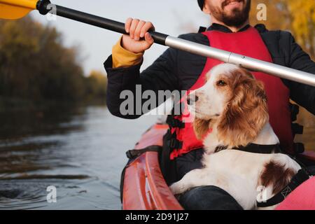 Mann rudert ein Kanu mit seinem Spaniel Hund, sonniges Herbstwetter. Kajakfahren mit Hunden auf dem Fluss, aktive Haustiere, glücklicher Hund und Besitzer auf einer Adove Stockfoto