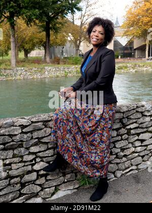 Vertikales Porträt einer attraktiven lächelnden afroamerikanischen Frau trägt Eine schwarze Jacke und sitzt an einem Fluss Stockfoto