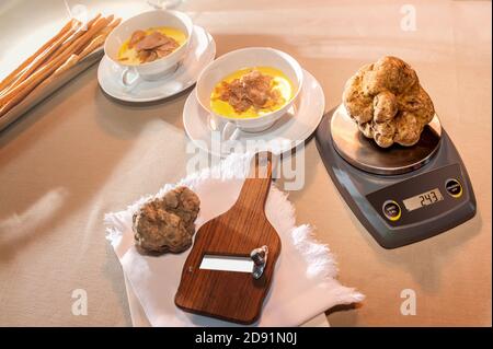 Waage mit einem großen schönen weißen Trüffel in der Nähe Holzschneidemaschine im Hintergrund zwei Tassen mit Käse Fondue und Trüffelscheiben, alle auf dem Tisch Stockfoto