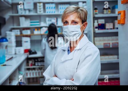 Senior Apotheker stehend suchen ernsthaft tragen Gesichtsmaske Schutz vor Coronavirus steht in der Apotheke Stockfoto