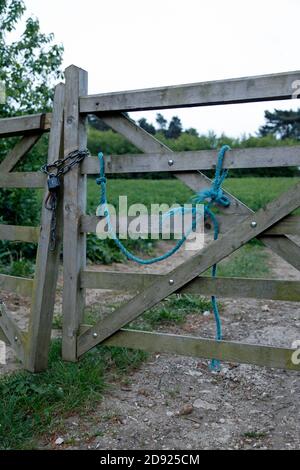 Ein 5-Stab-Holztor am Eingang zu einem Feld, kaum stehend und durch eine Kette und ein Zahlenschloss zusammengehalten. An einem Tor ist ein blaues Seil befestigt. Stockfoto