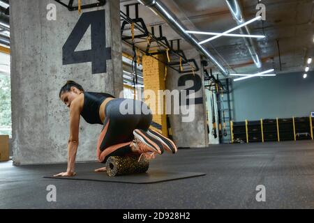 Rückansicht einer jungen, schönen Sportlerin in Sportbekleidung Training mit Schaumstoffrolle auf Yogamatte im Fitnessstudio, mit speziellen Fitnessgeräten. Sportliche Menschen, Training und Trainingskonzept Stockfoto