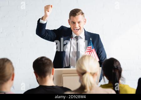 Aufgeregter Sprecher mit Gewinnergeste im Gespräch mit Rührern im Konferenzsaal, verschwommener Vordergrund Stockfoto