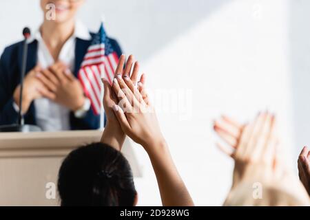 Dankbarer politischer Agitator, der die Hände auf der Brust hält, vor den Wählern, die im Konferenzraum applaudieren, verschwommener Vordergrund Stockfoto