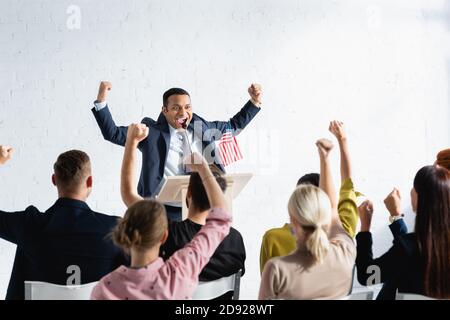 Aufgeregter indischer Sprecher, der eine Gewinnergeste zeigt, während er vor den Wählern im Konferenzsaal steht, verschwommener Vordergrund Stockfoto