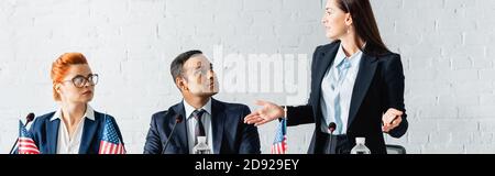 Politikerin mit offenen Armen und Blick auf multikulturelle Kollegen im Sitzungssaal, Banner Stockfoto