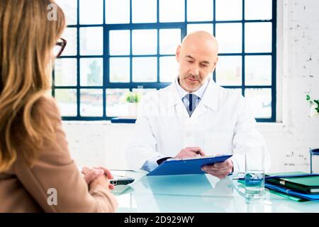 Schuss eines männlichen Arztes mittleren Alters, der mit seinem Patienten spricht, während er am Büroschreibtisch im Arztraum sitzt. Stockfoto