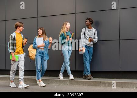 Lächelnde multiethnische Teenager mit Kaffee zum Gehen und Notebooks zu Fuß In der Nähe von Gebäuden im Freien Stockfoto