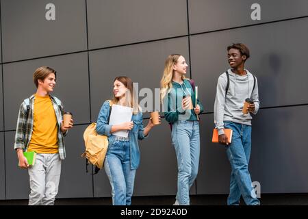 Positive multiethnische Teenager mit Rucksäcken und Kaffee zu Fuß gehen In der Nähe des Gebäudes Stockfoto