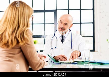 Schuss eines männlichen Arztes mittleren Alters, der mit seinem Patienten spricht, während er am Büroschreibtisch im Arztraum sitzt. Stockfoto