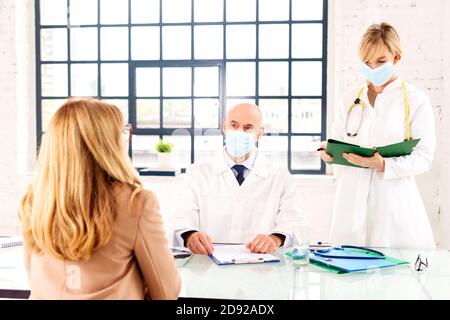 Aufnahme eines mittleren männlichen Arztes und einer Krankenschwester, die Gesichtsmaske trägt, während sie mit seinem Patienten im Zimmer des Arztes spricht. Stockfoto