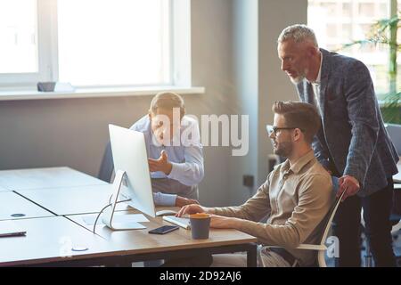 Drei männliche Coworker, die auf PC-Bildschirm schauen und über Projekt diskutieren, Geschäftsleute, die im Coworking Space zusammenarbeiten Stockfoto