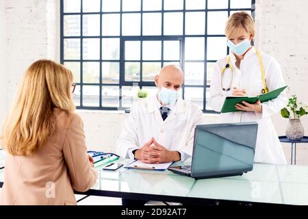 Aufnahme eines mittleren männlichen Arztes und einer Krankenschwester, die Gesichtsmaske trägt, während sie mit seinem Patienten im Zimmer des Arztes spricht. Stockfoto