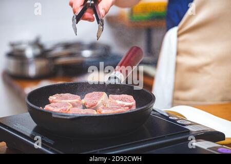 Fleisch in Speck wird in einer gusseisernen Pfanne gebraten, ein Mann Koch in einer Schürze dreht das Fleisch mit Metallzange in der Hand um. Küche, Lebensmittelzubereitung Stockfoto