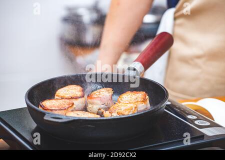 In Speck verpacktes Fleisch wird in einer gusseisernen Pfanne auf der Kochplatte gebraten, ein männlicher Koch trägt im Hintergrund eine Schürze. Küche, Essenszubereitung, kochen Stockfoto