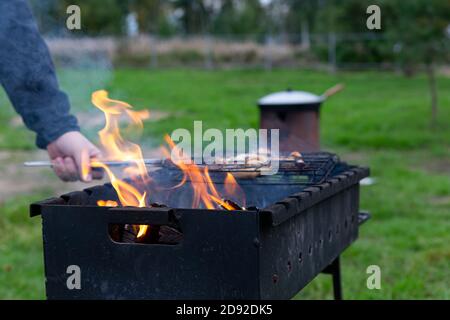 Das Fleisch wird auf dem Grill, Brazier gekocht. Nahaufnahme Mann die Hand dreht Fleisch auf dem Gitter über dem Feuer. Kochen im Freien. Treffen mit Freunden, Picknick, wir Stockfoto