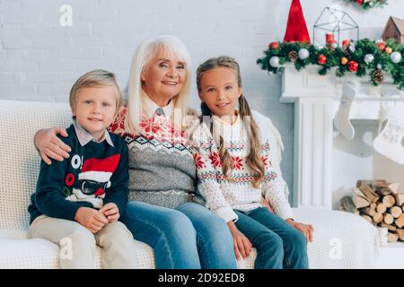 Glückliche ältere Großmutter umarmt Enkelkinder, während sie auf dem Sofa zusammen sitzt Stockfoto