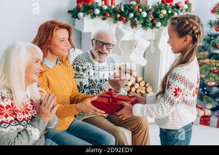 Lächelndes Mädchen präsentiert Geschenkbox zu glücklichen Mutter in der Nähe applaudieren Großeltern Stockfoto