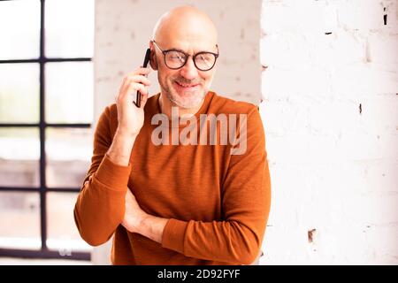 Porträtaufnahme eines glücklichen älteren Mannes in legerer Kleidung, der am Fenster steht und mit jemandem auf seinem Handy spricht. Stockfoto