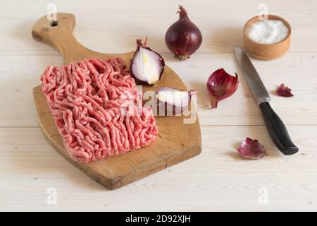 Hackfleisch und rote Zwiebeln liegen auf einem Holzbrett. Salzstreuer auf einem weißen Holztisch Stockfoto