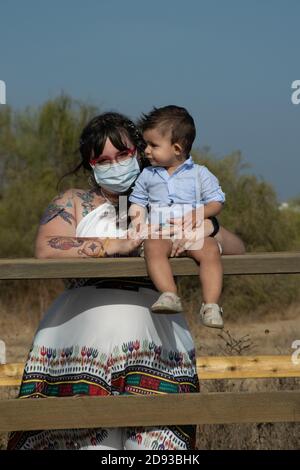 Kaukasische Frau mit Tätowierungen auf ihrem Arm trägt eine Gesichtsmask Während sie ihr Kind auf einem Holz sitzen lassen Stockfoto