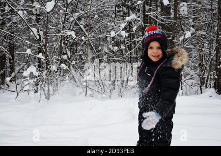 Ein schlichtes kleines Kind wirft Schnee im Winter schneebedeckten Wald, Bäume im Schnee. Genießt den Winter. Spielt Schneebälle. Glücklich, lächelnd, lacht Stockfoto