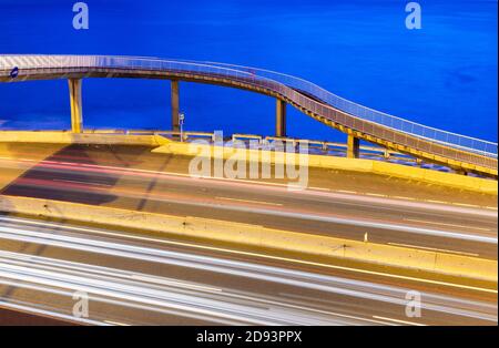 Lange Exposition von Lichtspuren aus dem Verkehr auf Küstenstraße mit Fußgängerbrücke und Meer im Hintergrund bei Sonnenaufgang. Stockfoto