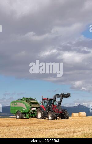 Ein Red Valtra Traktor und McHale Fusion Vario Ballenpresse in Betrieb In Aberdeenshire mit Bennachie Visible in The Distance Stockfoto