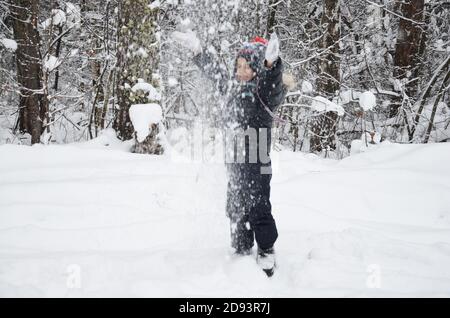 Ein schlichtes kleines Kind wirft Schnee im Winter schneebedeckten Wald, Bäume im Schnee. Genießt den Winter. Spielt Schneebälle. Glücklich, lächelnd, lacht Stockfoto