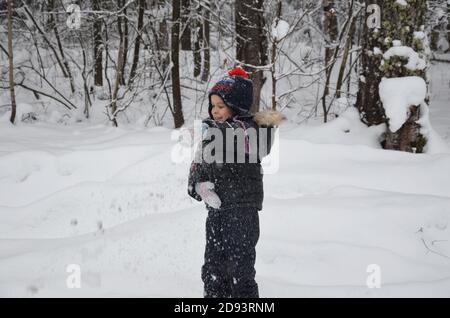 Ein schlichtes kleines Kind wirft Schnee im Winter schneebedeckten Wald, Bäume im Schnee. Genießt den Winter. Spielt Schneebälle. Glücklich, lächelnd, lacht Stockfoto