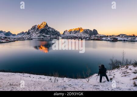 Landschaftsfotograf mit Stativ von hinten in der Nähe der kleinen gesehen Fischerdorf reine auf den Lofoten Inseln in Norwegen Winter mit steiler schneekappe Stockfoto