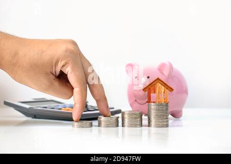 Die Hände des männlichen Investors werden auf den Münzstapel gelegt, einschließlich des Holzhausmodells auf dem Münzstapel, Geldsparideen, Unternehmenswachstum und Inve Stockfoto