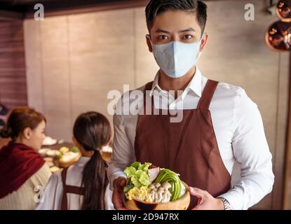 Junger Kellner trägt eine schützende Gesichtsmaske, während er Essen serviert Kunde im Restaurant Stockfoto