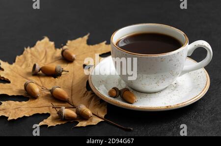Stillleben im Herbst mit einer Tasse Kaffee, Blättern und Eicheln auf schwarzem Hintergrund. Acorn-Kaffee. Koffein-Ersatz. Stockfoto