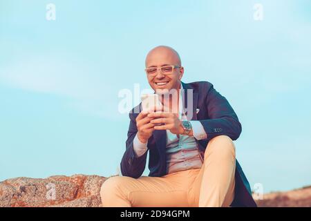 Glücklicher kahlköpfiger Geschäftsmann lächelnder Mann, der auf dem Mobiltelefon auf Felsen im Freien am blauen Himmel auf dem Hintergrund sitzt. Positive Gesichtsausdruck menschliche Emotion r Stockfoto