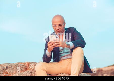 Nahaufnahme Porträt Kopfschuss Mann mittleren Alters mit Brille unglücklich skeptisch Blick auf den Handy-Bildschirm draußen sitzen auf Felsen Blauer Himmel Hintergrund Stockfoto