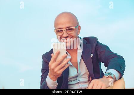 Glücklicher kahlköpfiger Geschäftsmann lächelnder Mann, der auf dem Mobiltelefon auf Felsen im Freien am blauen Himmel auf dem Hintergrund sitzt. Positive Gesichtsausdruck menschliche Emotion r Stockfoto