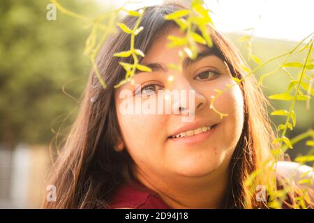 Portrait der hispanischen Frau bei Sonnenuntergang umgeben von grünem Baum Zweige mit Sonnenlicht dahinter Stockfoto