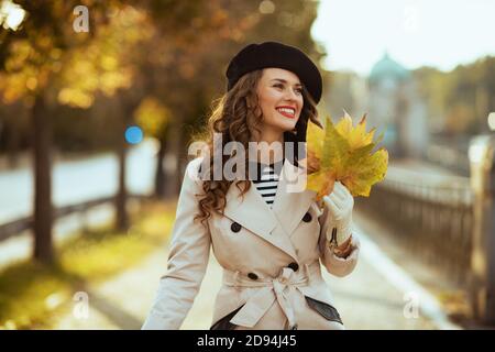 Hallo Herbst. Glücklich elegante 40 Jahre alte Frau in beige Trenchcoat und schwarze Baskenmütze mit Herbst gelbe Blätter im Freien in der Stadt im Herbst. Stockfoto