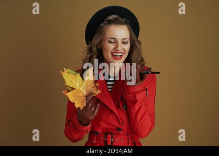 Hallo oktober. Lächelnd stilvolle 40 Jahre alte Frau in rotem Mantel und schwarzer Baskenmütze mit Smartphone, Lederhandschuhe und gelben Herbst Ahornblätter auf beige Stockfoto