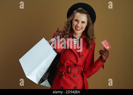 Hallo september. Porträt von lächelnd stilvolle 40 Jahre alte Frau in rotem Mantel und schwarze Baskenmütze mit Kreditkarte, Lederhandschuhe und Papier Einkaufstaschen i Stockfoto