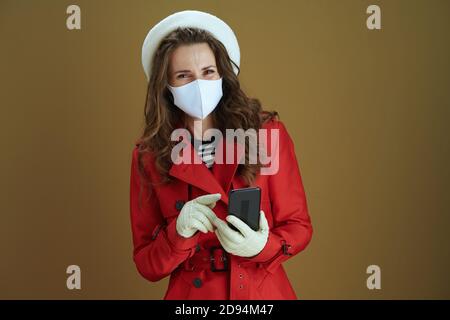 Hallo Herbst. Lächelnd elegante 40 Jahre alte Hausfrau in rotem Mantel mit Smartphone-App vor braunem Hintergrund. Stockfoto